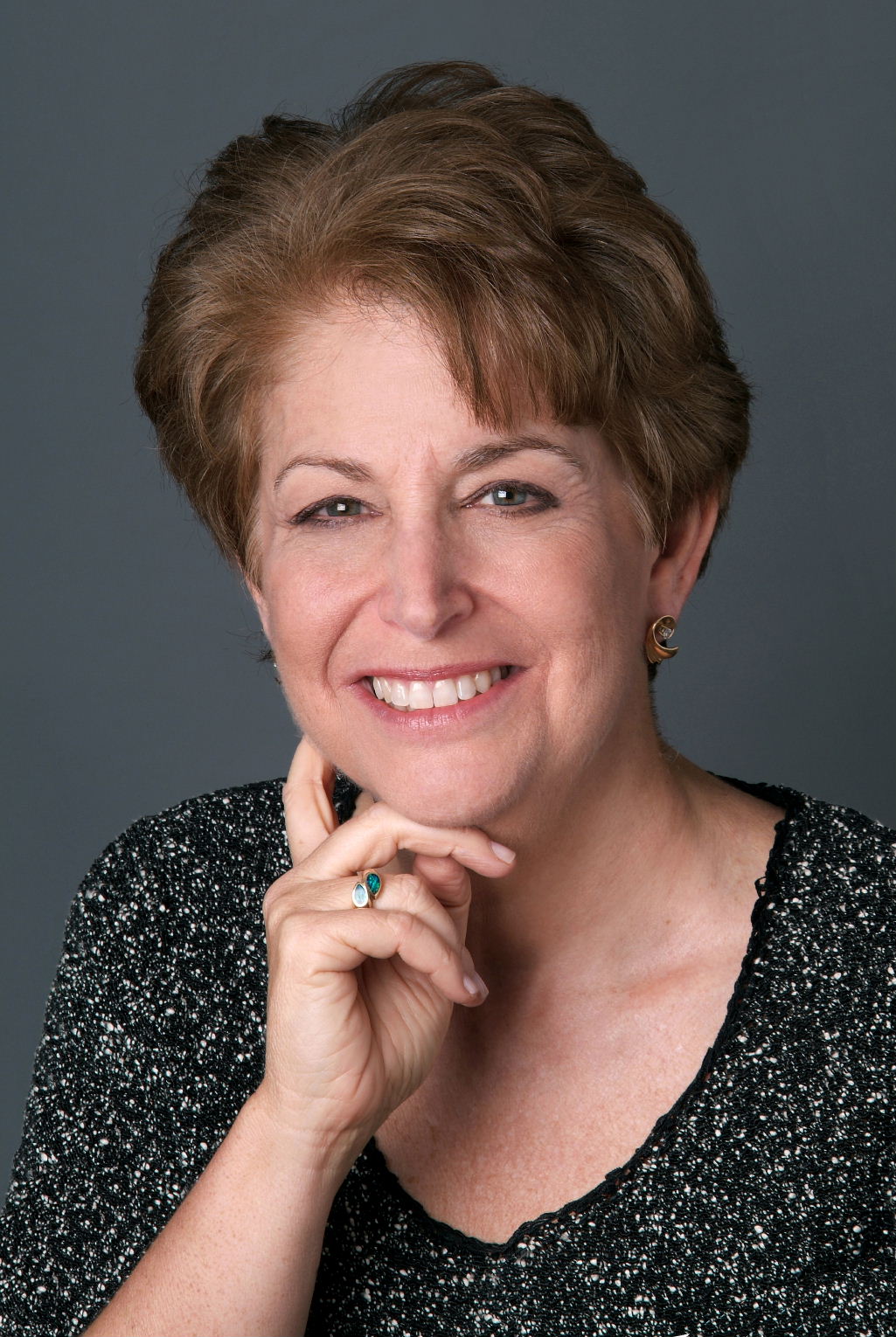 A portrait of Dr. Diane with short, brown wavy hair, smiling with her right hand on her right cheek. She is wearing a black and white speckled, v-neck blouse.