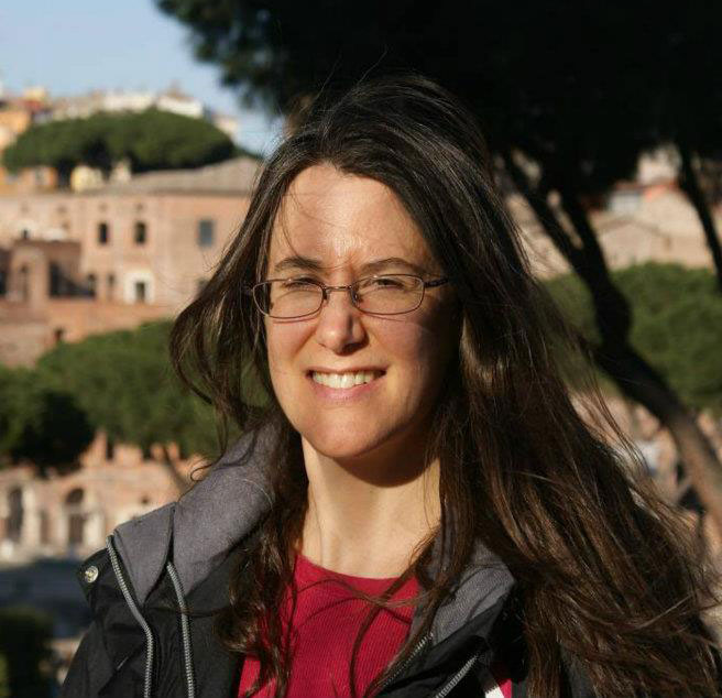 A portrait of Clara Diebold smiling outside in the sun with a large tree and pink stone buildings behind her. She is wearing a casual red shirt under a gray coat, glasses and has long brown hair.