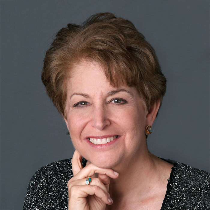 A portrait of Dr. Diane with short, brown wavy hair, smiling with her right hand on her right cheek. She is wearing a black and white speckled, v-neck blouse.