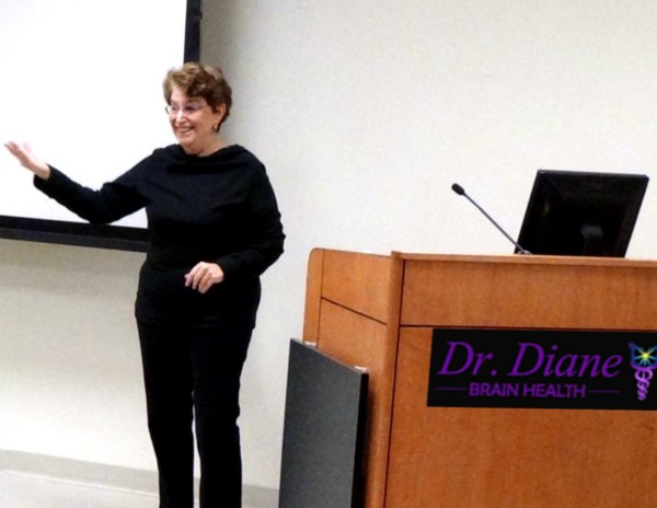 A picture of Dr. Diane speaking as an inspirational professional speaker. She is wearing a black shirt and black pants and holds her arm out and smiling. She has short brown wavy hair and stands to the left of a podium with her name. 