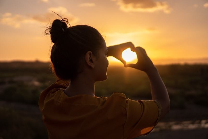 Woman looking at the sunrise through her hands shaped like a heart.